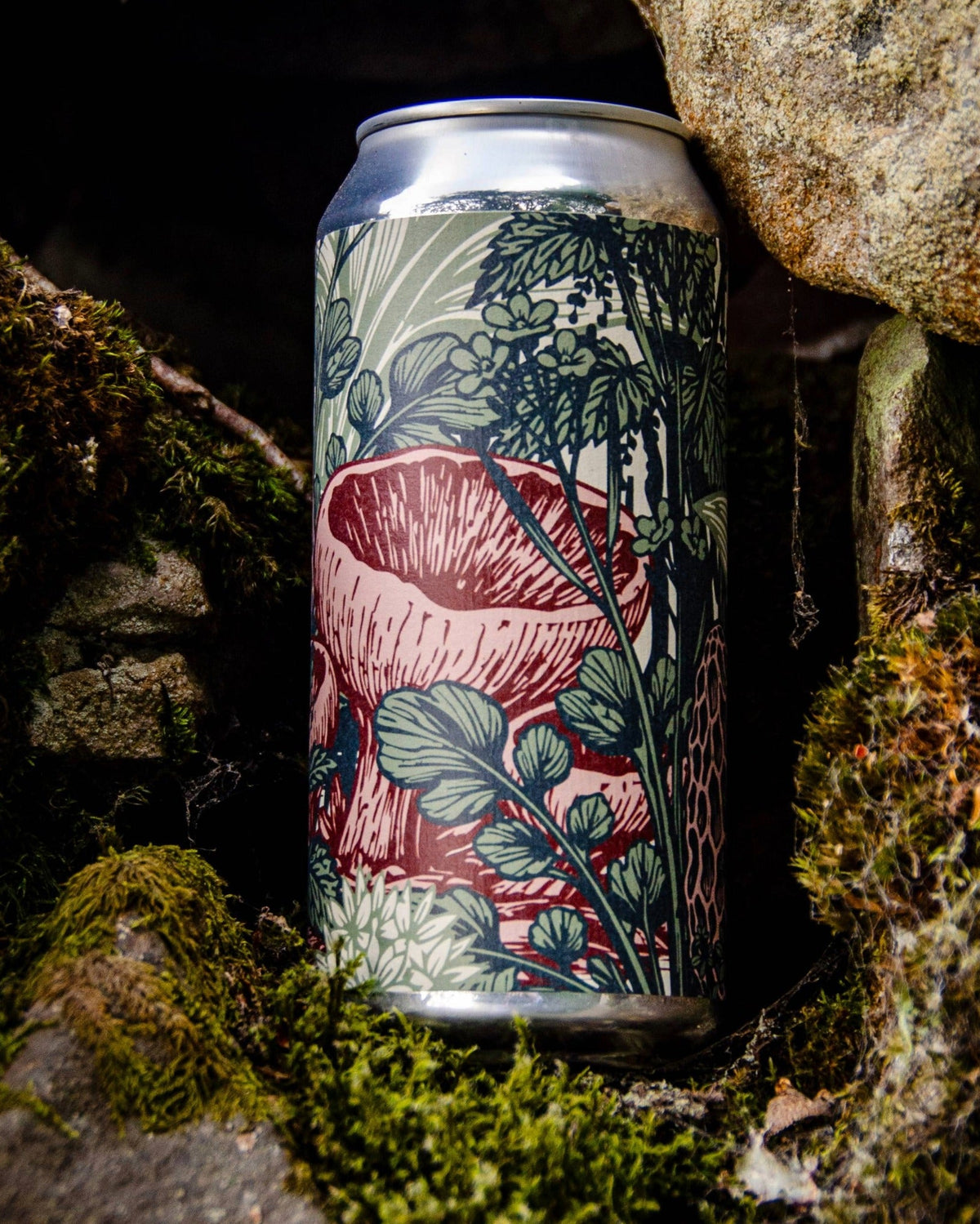 A slim aluminum craft beer can with a detailed botanical mushroom illustration, set in a natural mossy rock environment.